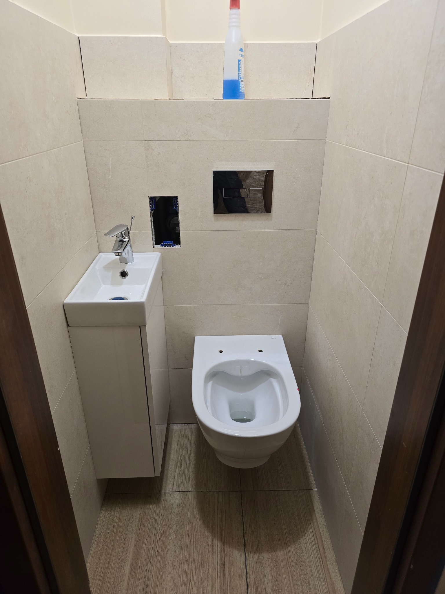 Wykończona, wąska łazienka w Kożuchowie z beżowymi płytkami, wiszącą toaletą i małą umywalką z szafką. Widoczne przygotowane otwory na armaturę.