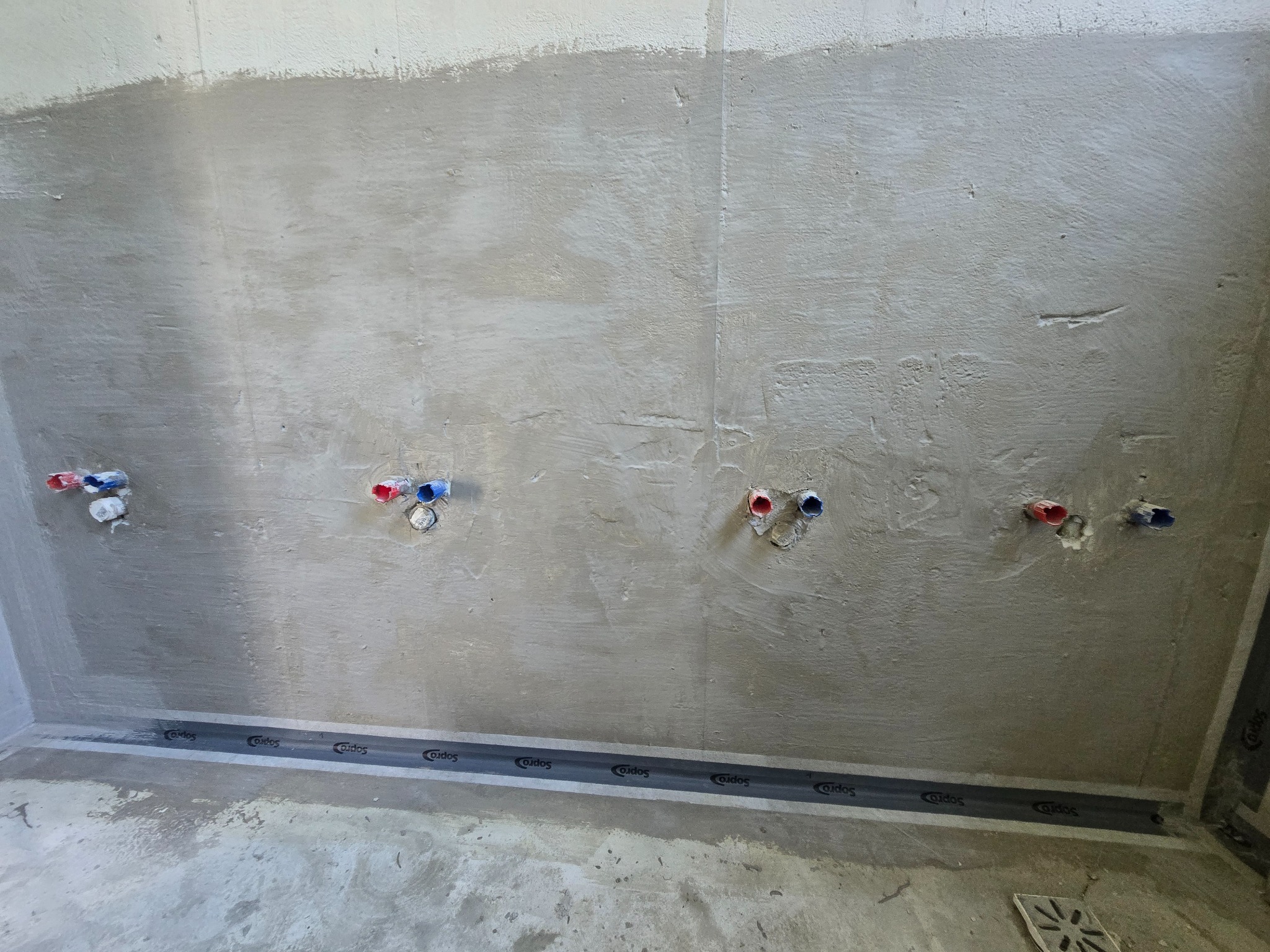 Przygotowana ściana w łazience z wyprowadzonymi instalacjami wodnymi i zamontowanym odpływem liniowym wzdłuż ściany. Ściana zagruntowana, gotowa do dalszych prac wykończeniowych.