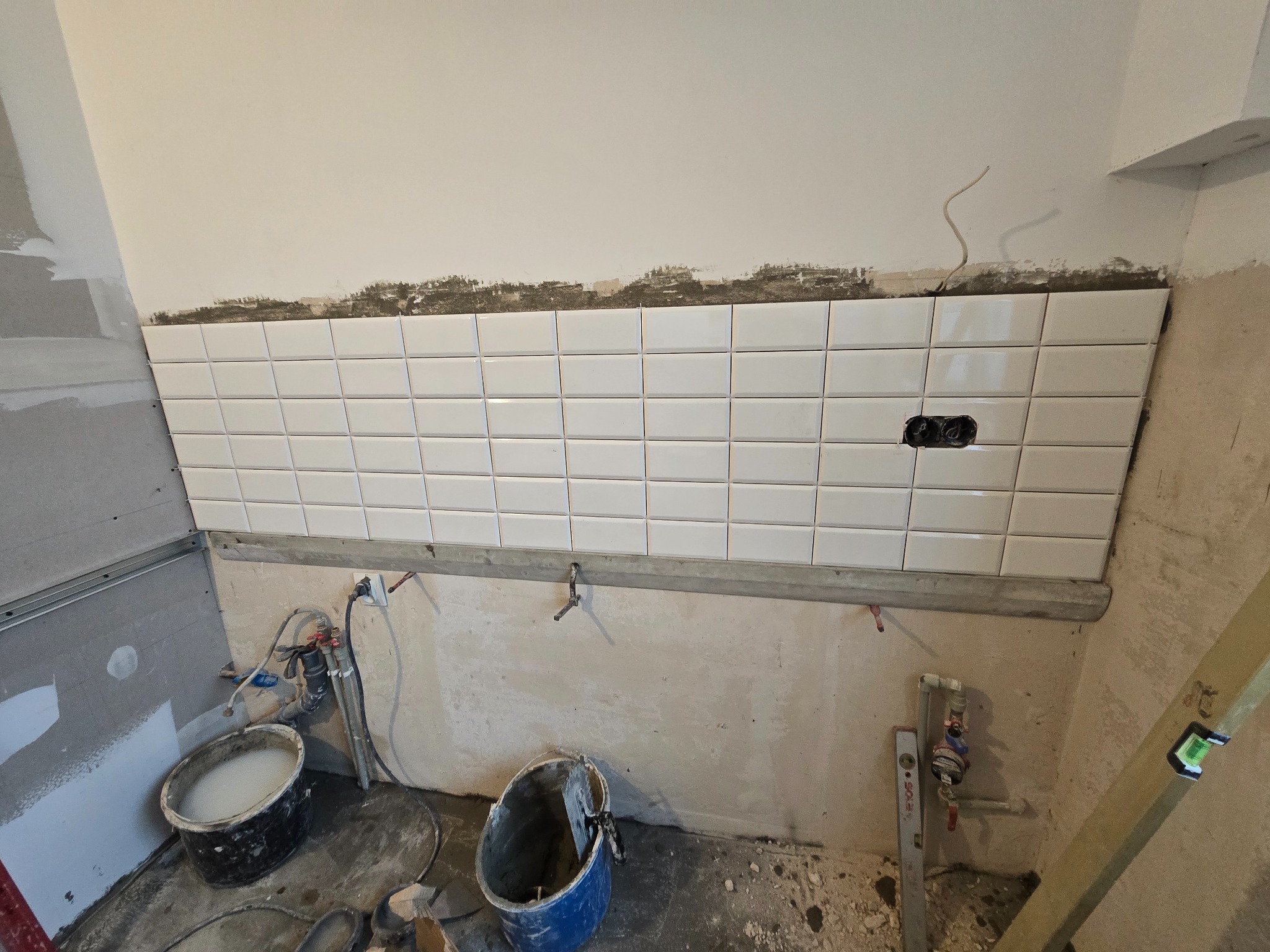 Białe, prostokątne płytki ułożone na ścianie w trakcie remontu. Widoczne narzędzia, wiadro z zaprawą i poziomica. Fragment instalacji elektrycznej.
