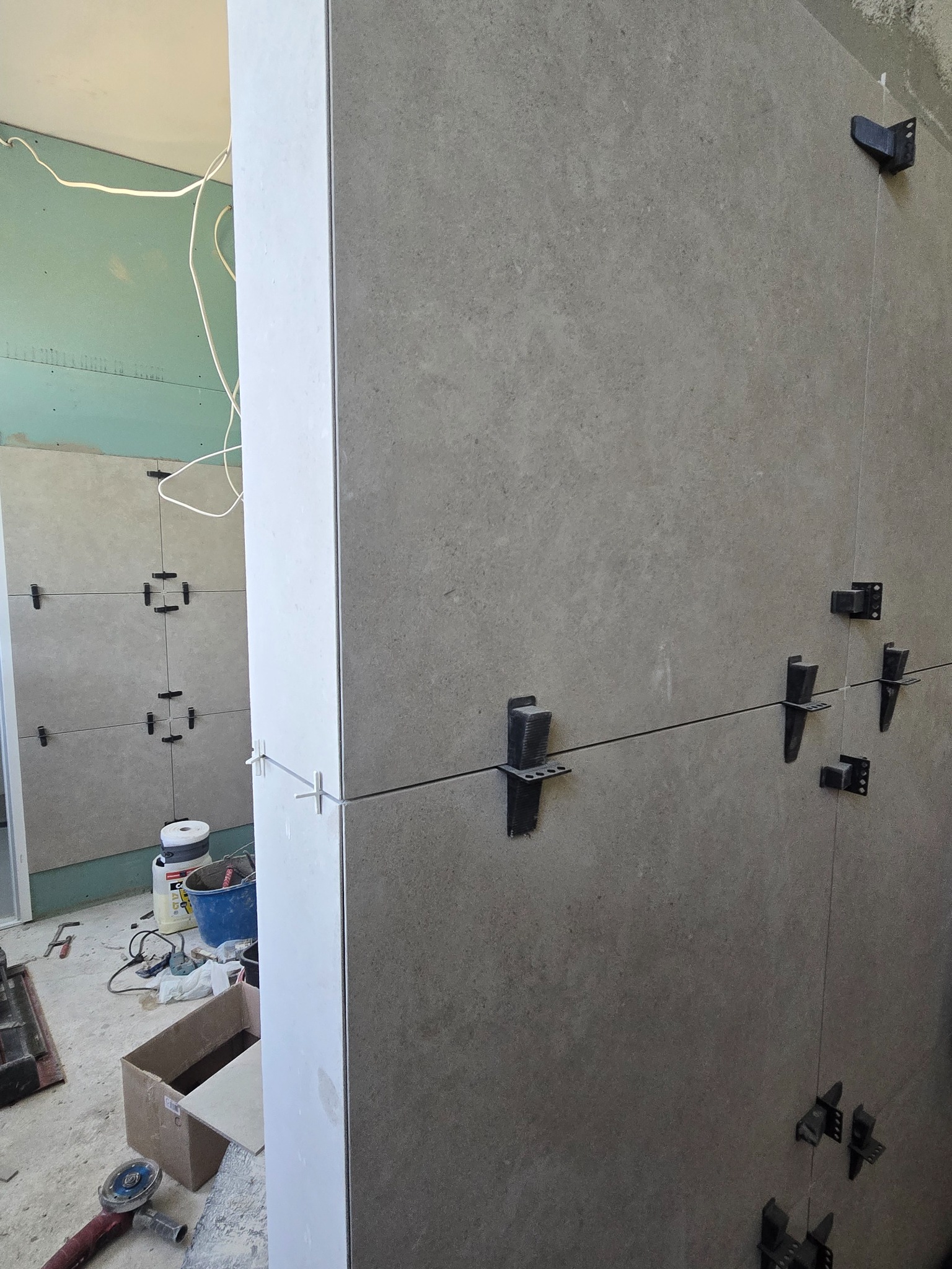 Montaż płytek ściennych z użyciem klipsów poziomujących. Widoczne narzędzia i materiały budowlane. Szare płytki o jednolitej fakturze. Prace remontowe w toku.