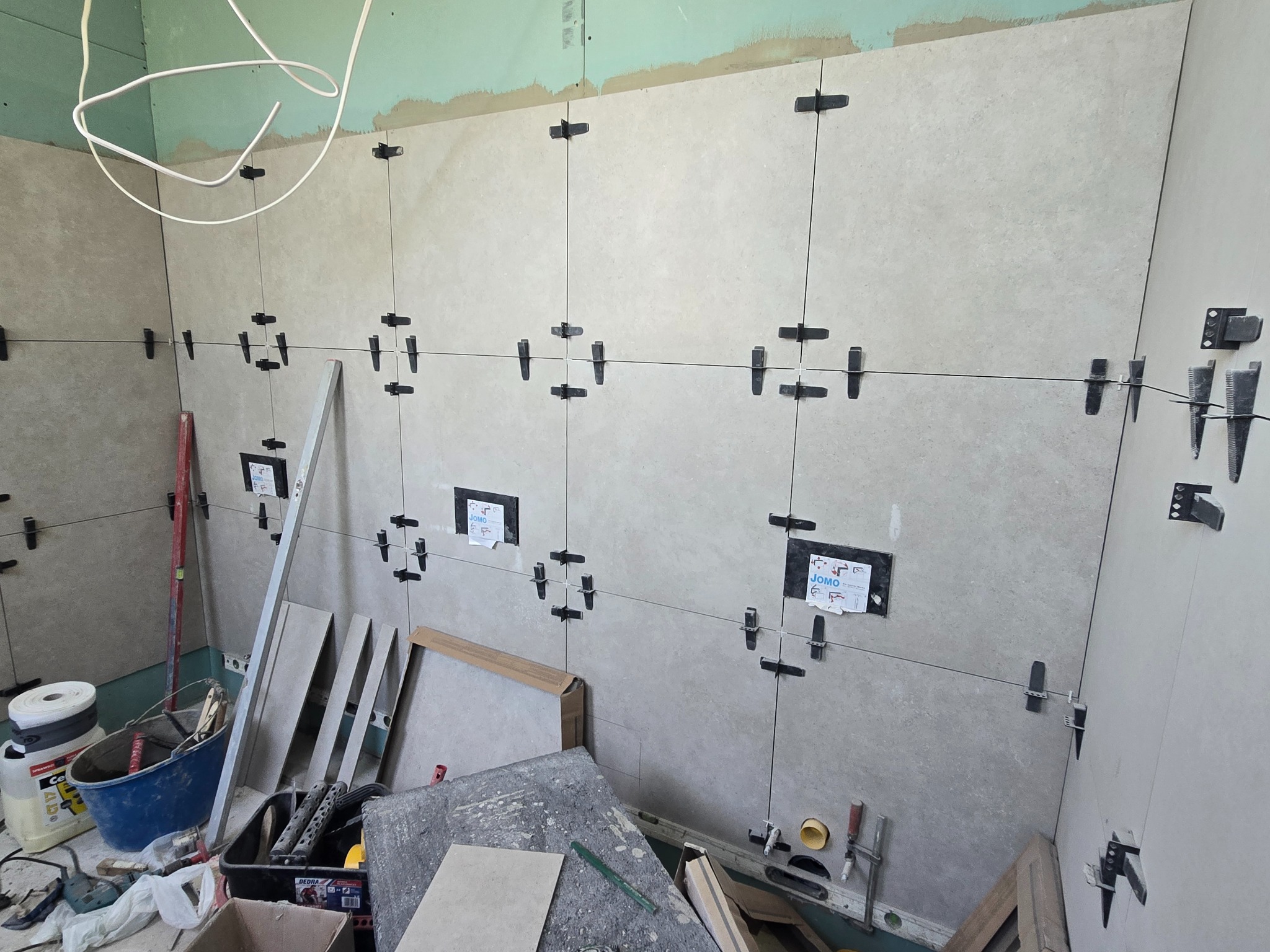 Ściana w trakcie układania płytek z użyciem systemu poziomowania. Widoczne narzędzia i materiały budowlane. Prace remontowe w łazience.