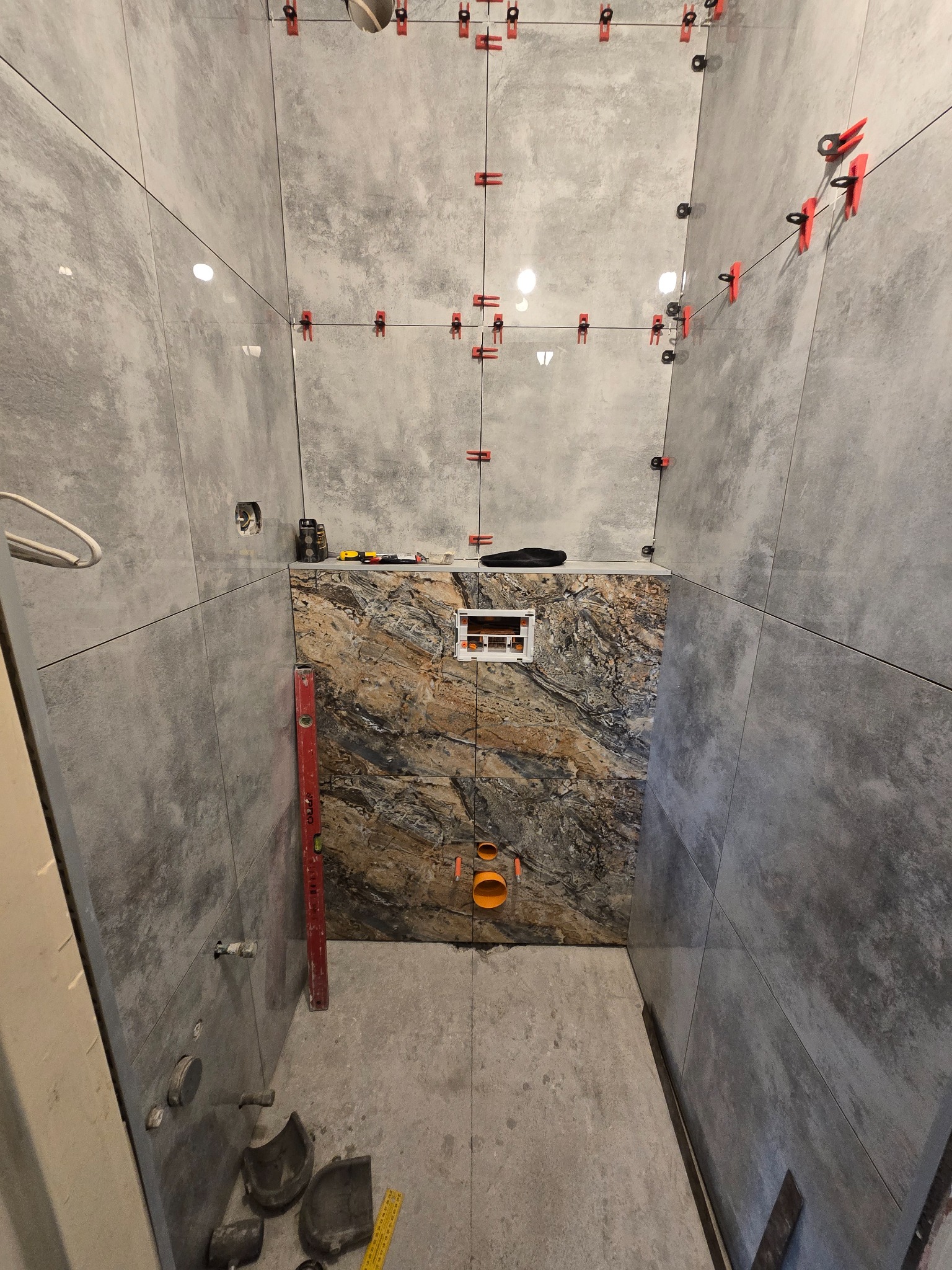 Wnętrze łazienki w trakcie remontu: szare płytki na ścianach z czerwonymi klipsami poziomującymi, fragment ściany z dekoracyjną płytką, poziomica i narzędzia na podłodze.