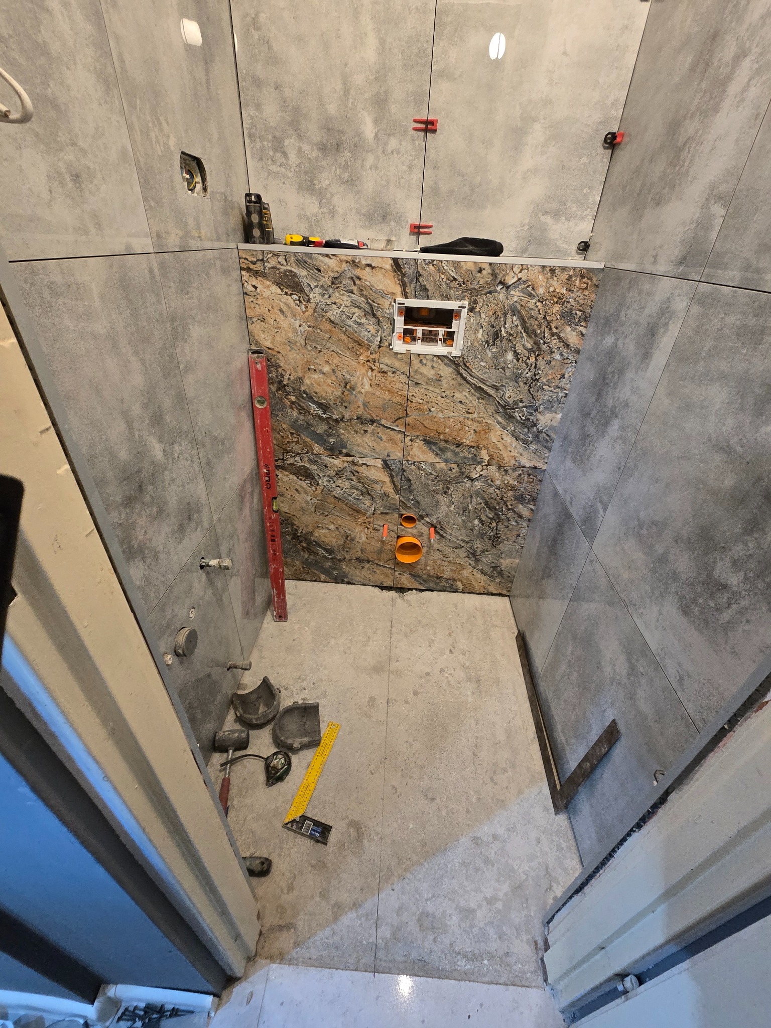 Wnętrze łazienki w trakcie remontu: szare płytki ścienne, fragment ściany z dekoracyjną okładziną, widoczne narzędzia i elementy instalacji. Prace wykończeniowe w toku.