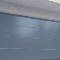 Szara, segmentowa brama garażowa z poziomymi przetłoczeniami, widoczna z bliska, z żółtą naklejką w prawym górnym rogu i fragmentem folii ochronnej w prawym dolnym rogu.