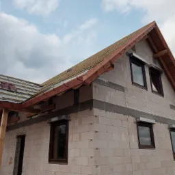 Budowa domu jednorodzinnego z zamontowanymi oknami i przygotowaniem pod rolety zewnętrzne, widoczne drewniane elementy konstrukcji dachu oraz białe ściany z betonu komórkowego.