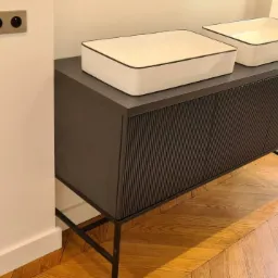 Nowoczesna szafka łazienkowa w kolorze grafitowym z ryflowanymi frontami, na metalowych nogach, z dwiema białymi umywalkami nablatowymi, na parkiecie ułożonym w jodełkę.