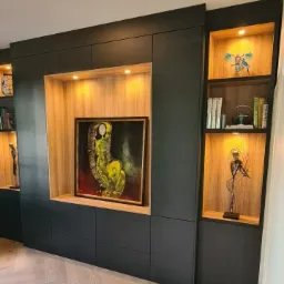 Ciemnogranatowa zabudowa meblowa z oświetlonymi wnękami, w jednej z nich obraz w złotej ramie, w pozostałych dekoracje i książki, parkiet w jodełkę na podłodze.