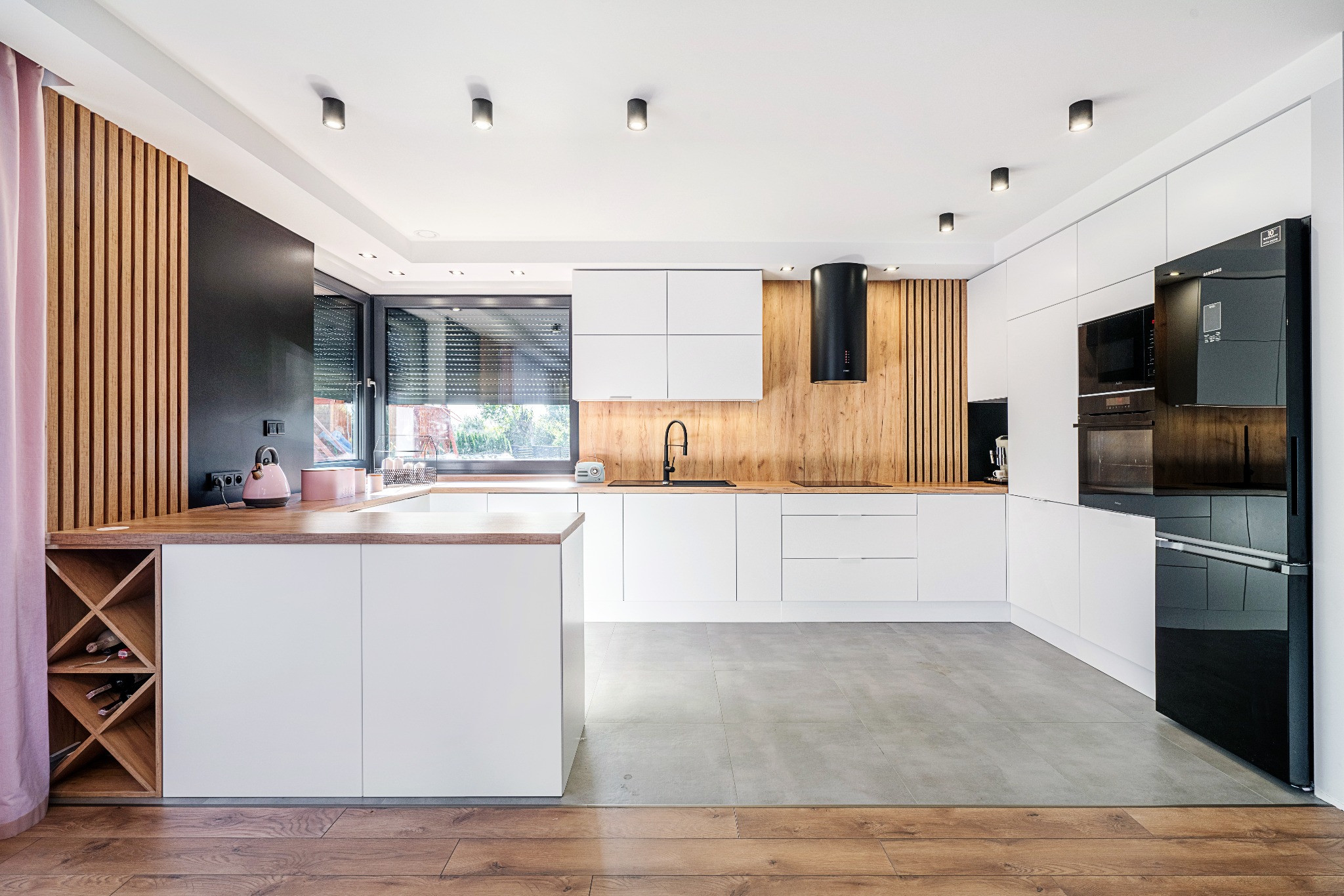 Nowoczesna kuchnia z białymi szafkami, drewnianym blatem i akcentami w kolorze drewna, czarnym okapem i lodówką, oraz wyspą z regałem na wino.