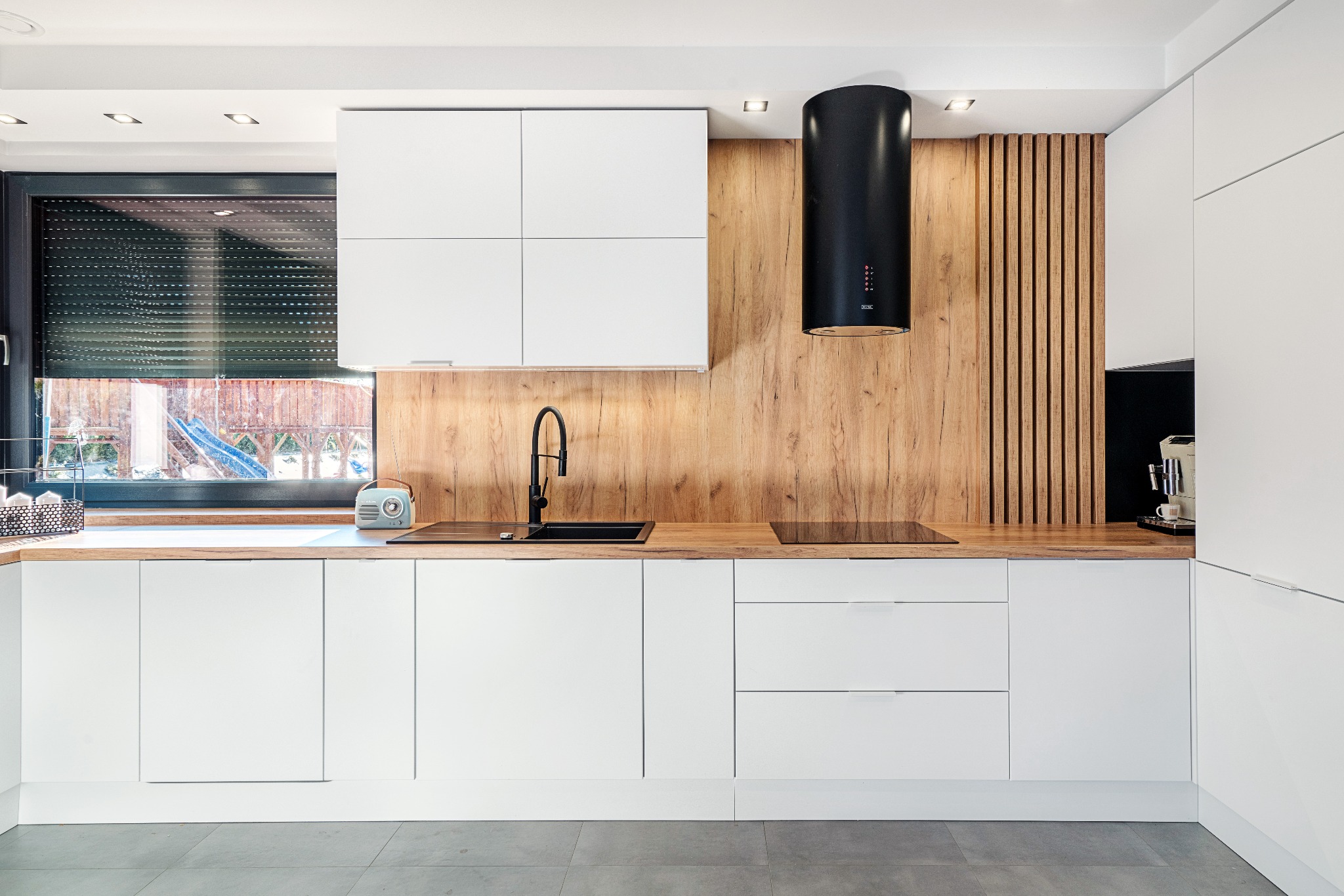 Nowoczesna kuchnia z białymi frontami, drewnianym blatem i okładziną ścienną, czarny okap cylindryczny, bateria i zlew, retro radio na blacie.