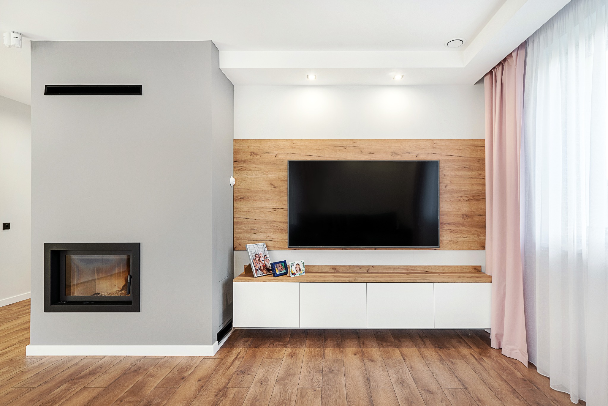 Nowoczesny salon z drewnianą podłogą, szarą ścianą z kominkiem, szafką RTV z drewnianym blatem i białymi frontami, telewizorem, trzema ramkami ze zdjęciami oraz delikatnymi różowymi zasłonami.