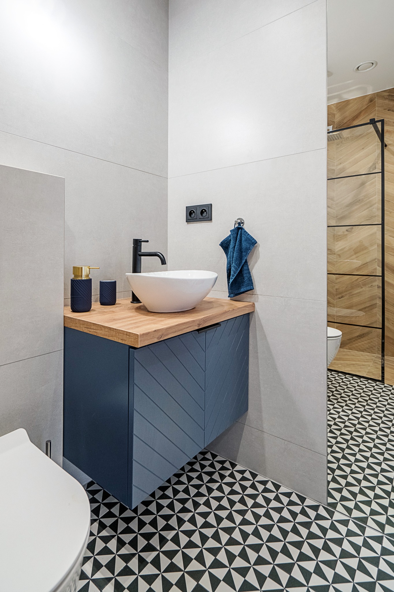 Nowoczesna łazienka z umywalką nablatową na drewnianym blacie, szafka w kolorze granatowym z frezowanymi frontami, wzorzysta podłoga i kabina prysznicowa z drewnopodobnymi płytkami w tle.