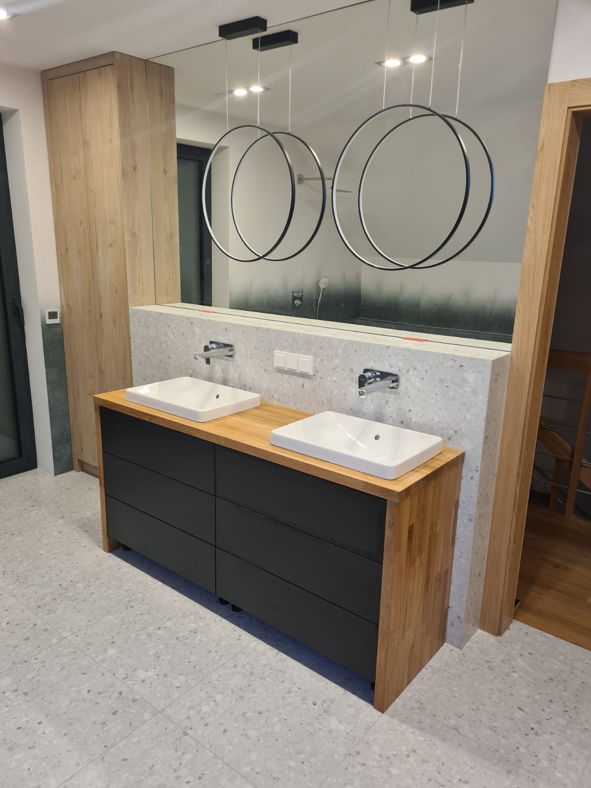 Nowoczesna łazienka z podwójną umywalką na drewnianym blacie z czarnymi szufladami, duże lustro z okrągłymi lampami wiszącymi.
