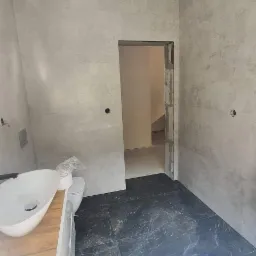 Wykończona łazienka z szarymi płytkami imitującymi beton na ścianach, ciemną marmurkową podłogą, wiszącą umywalką na drewnianej szafce i nowoczesną lampą sufitową z kulistymi kloszami.