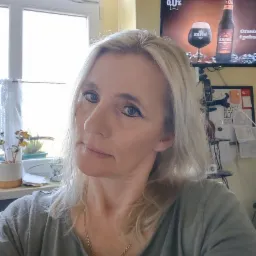 Portret kobiety z blond włosami, w tle okno z firanką i roślina w doniczce oraz telewizor z reklamą piwa Karmi.