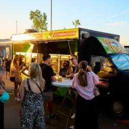 Food truck 'Bomba Truck Ł&oacute;dź' serwujący burgery. Klienci stoją przy stoliku, na zewnątrz, w słoneczny dzień. Widoczne menu z burgerami.