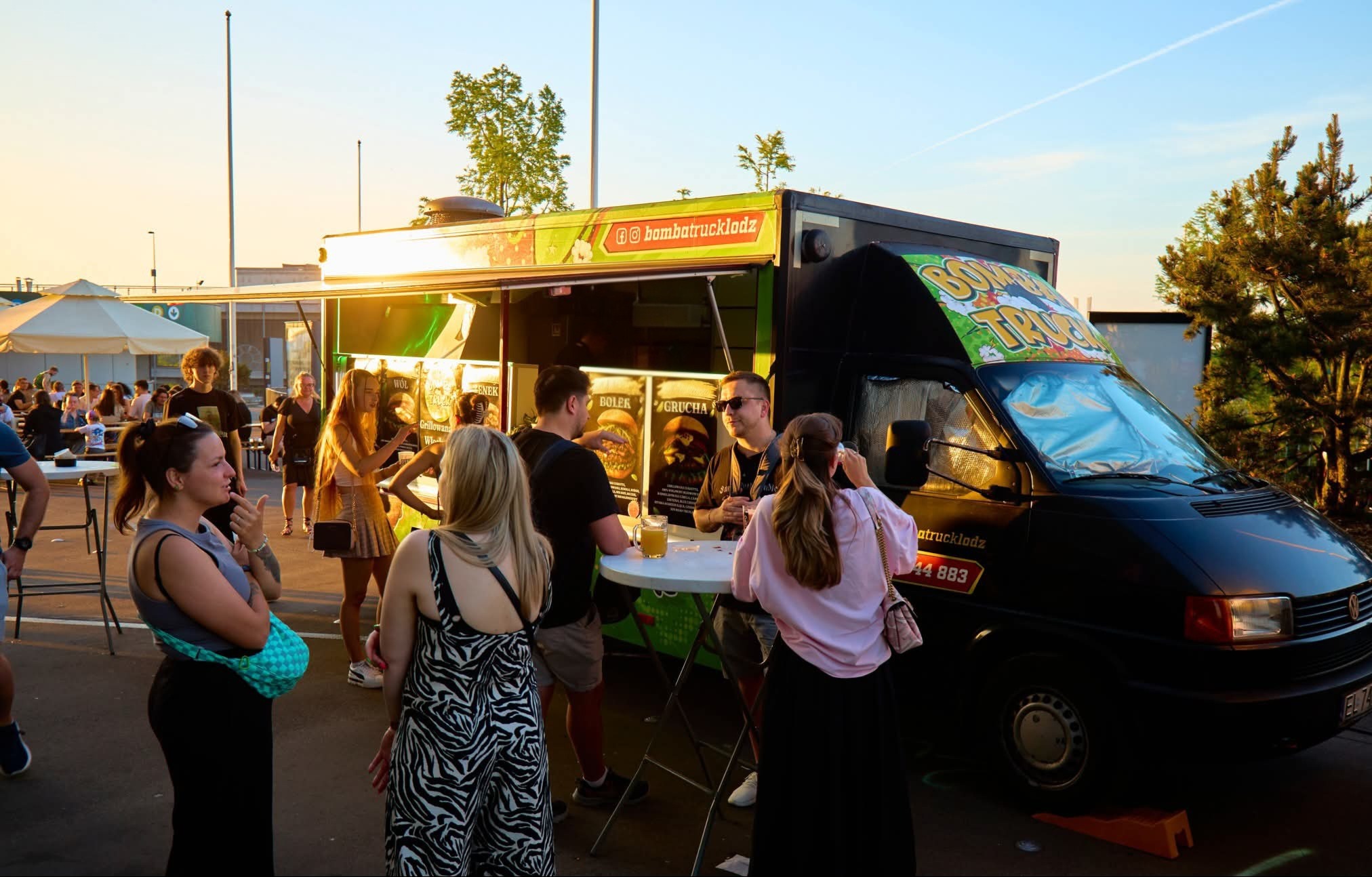 Food truck 'Bomba Truck Łódź' serwujący burgery. Klienci stoją przy stoliku, na zewnątrz, w słoneczny dzień. Widoczne menu z burgerami.