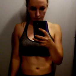 Kobieta w sportowym staniku i legginsach robi selfie w lustrze, widoczny telefon w dłoni.