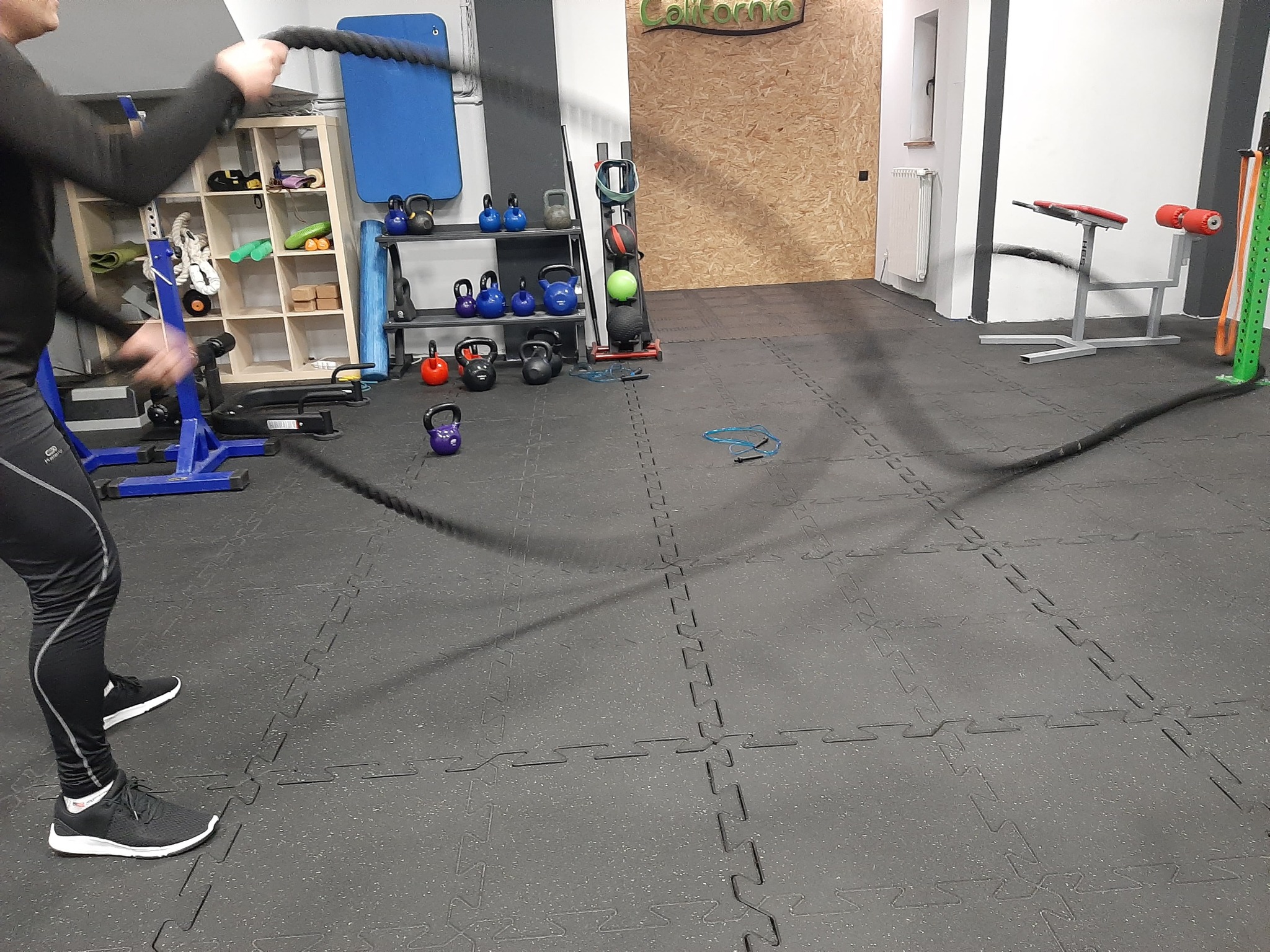 Osoba w czarnym stroju sportowym ćwiczy z grubymi linami w siłowni wyposażonej w kettlebells i inne przyrządy do ćwiczeń.
