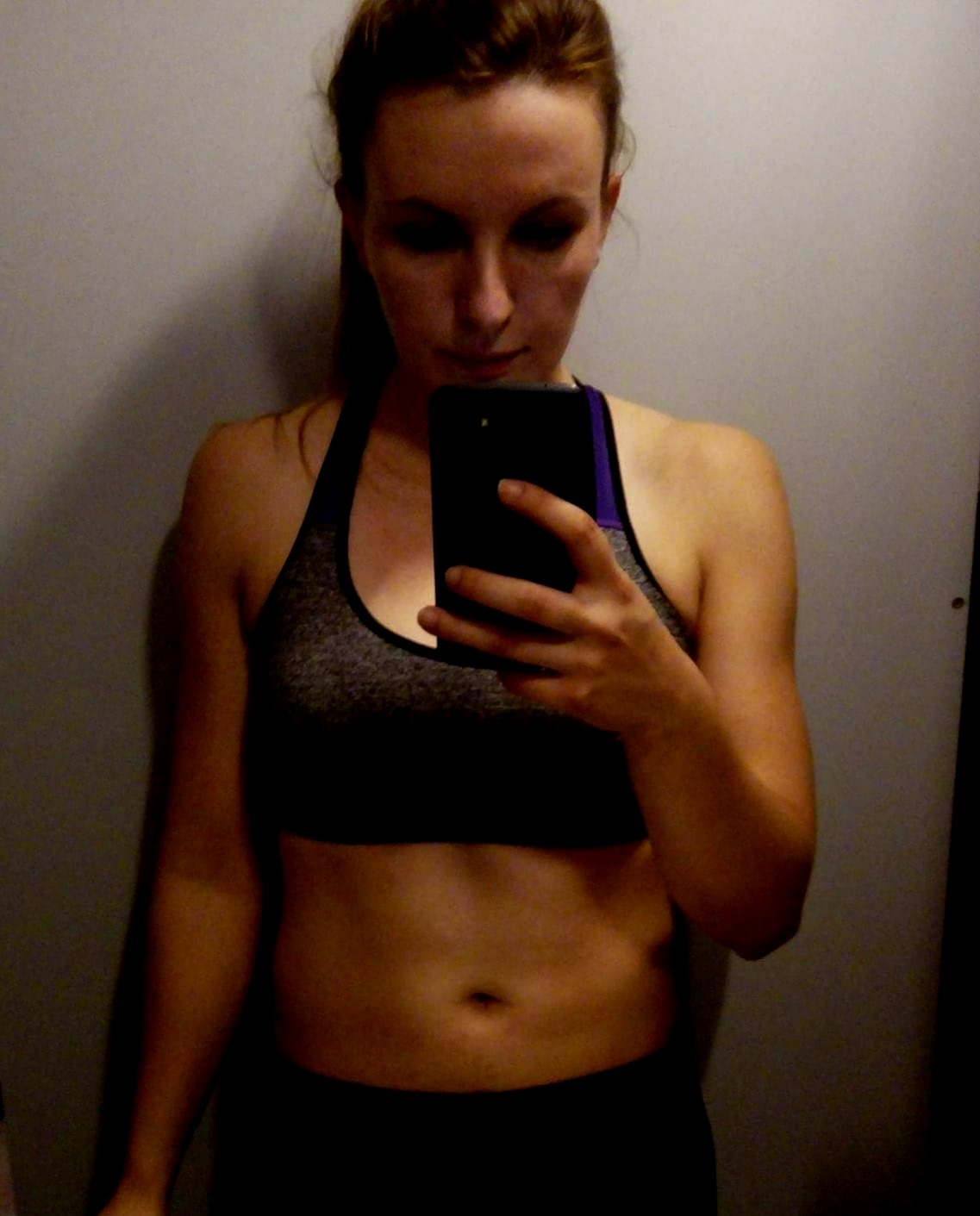 Kobieta w sportowym staniku i legginsach robi selfie w lustrze, widoczny telefon w dłoni.