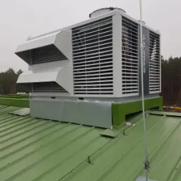 Duża centrala wentylacyjna Flowair z lamelami i dwoma daszkami na dachu pokrytym zieloną blachą trapezową, widoczny maszt odgromowy i las w tle.