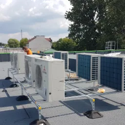 Instalacja szeregu klimatyzatorów Mitsubishi na dachu budynku, widoczny monter w pomarańczowej koszulce, system wsporników z żółtymi elementami, tło z budynkami i drzewami, pochmurne niebo.