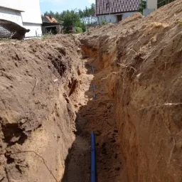 Wąski wykop w ziemi z ułożonymi niebieskimi rurami, widoczne ściany wykopu z różnymi warstwami gleby, w tle budynki mieszkalne i drzewa pod błękitnym niebem.