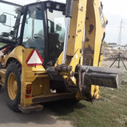 Żółta koparko-ładowarka CAT z otwartymi drzwiami kabiny stoi na szutrowej drodze, w tle hałda gruzu i słup wysokiego napięcia.