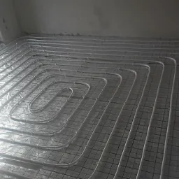 Białe rury grzewcze ułożone spiralnie na metalowej siatce, przygotowane do zalania wylewką, widok z góry na tle surowych ścian i okna.