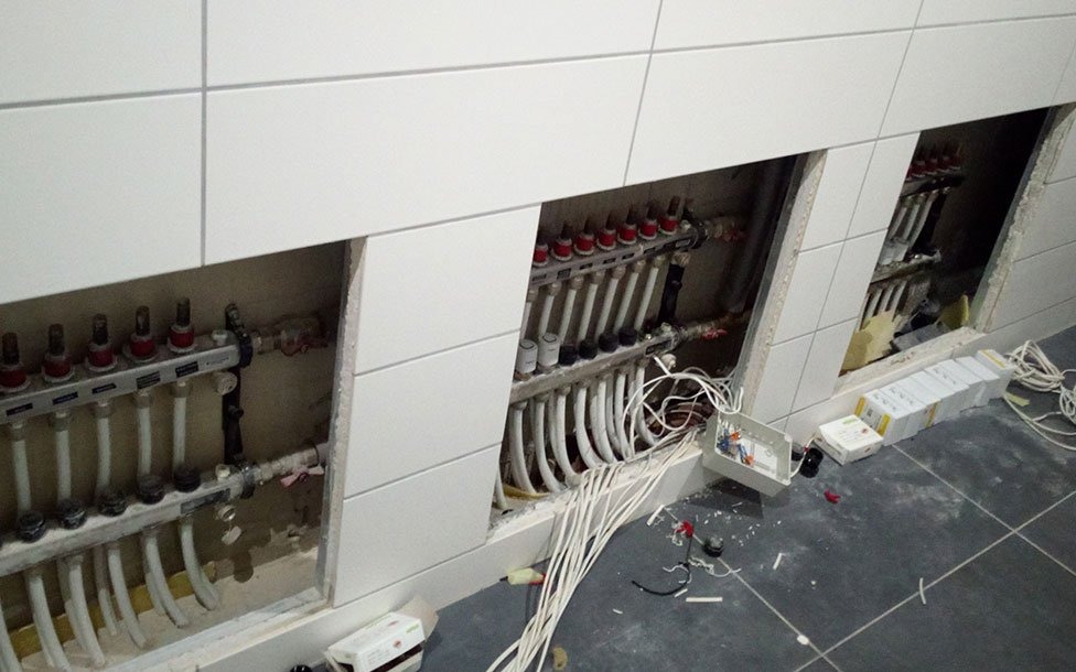 Rozdzielacze instalacji grzewczej podtynkowe, z widocznymi rurami i przewodami elektrycznymi, w trakcie montażu w pomieszczeniu z wykończonymi płytkami ściennymi i podłogowymi.