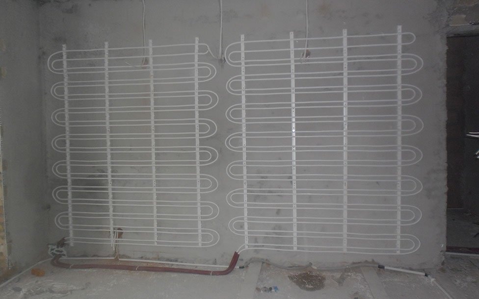 Białe rury systemu ogrzewania podłogowego zamontowane na szarej ścianie w surowym stanie, widoczne również rury doprowadzające.