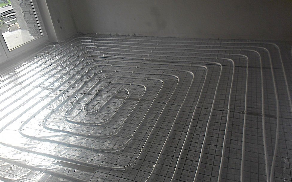 Białe rury grzewcze ułożone spiralnie na metalowej siatce, przygotowane do zalania wylewką, widok z góry na tle surowych ścian i okna.