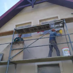 Dwóch pracowników na rusztowaniu podczas natryskowego nakładania tynku na elewację domu, z widocznym wiadrem farby Lakma-Tex.