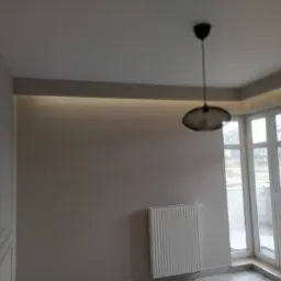 Wnętrze pokoju po remoncie z grzejnikiem, dużym oknem balkonowym i nowoczesną lampą wiszącą. Ściany pomalowane na szaro, widoczne subtelne listwy dekoracyjne. Na suficie zamontowane oświetlenie...