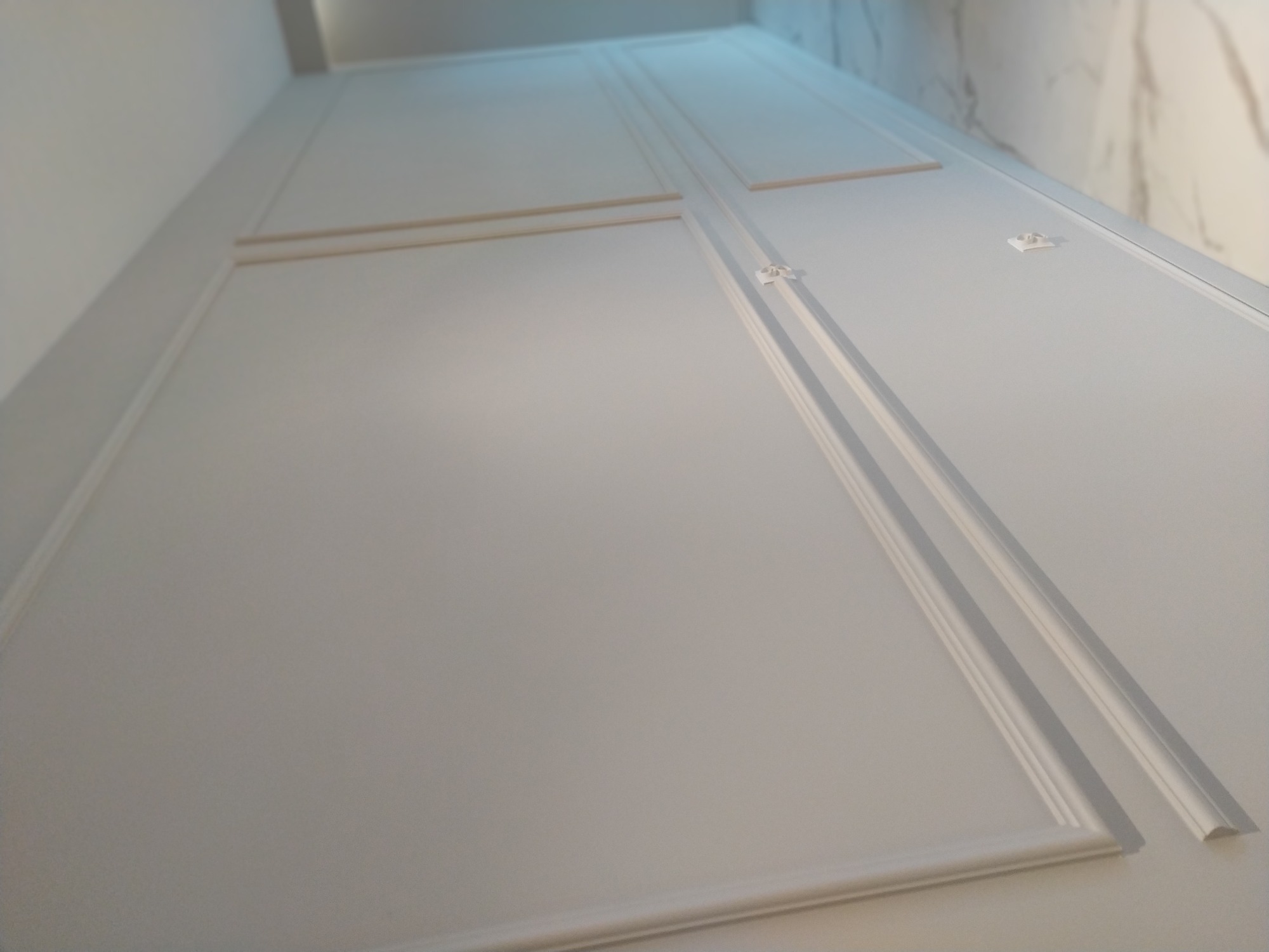 Białe panele ścienne z dekoracyjnymi listwami i ozdobnymi elementami łączącymi, zamontowane na ścianie z efektem marmuru w tle.