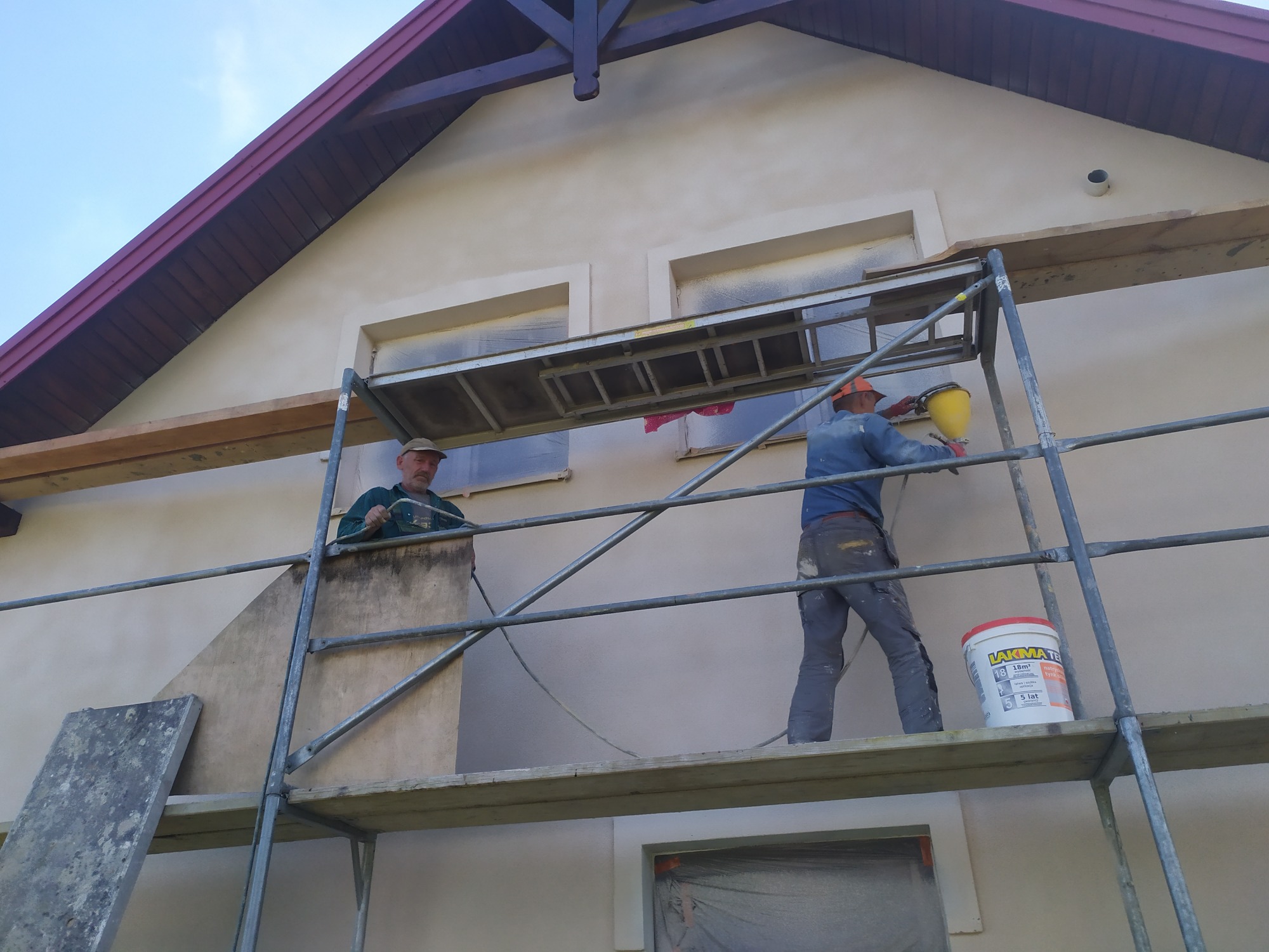 Dwóch pracowników na rusztowaniu podczas natryskowego nakładania tynku na elewację domu, z widocznym wiadrem farby Lakma-Tex.