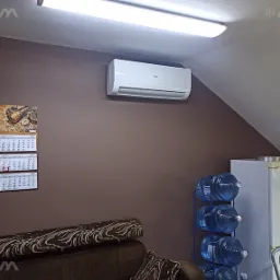 Biały klimatyzator Haier zamontowany na brązowej ścianie w pokoju na poddaszu, obok kalendarza i regału z butlami wody.