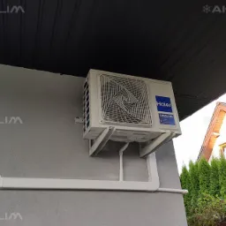 Zewnętrzna jednostka klimatyzatora Haier zamontowana na szarej ścianie domu z widocznymi białymi rurami odprowadzającymi, na tle zielonych drzew i fragmentu dachu sąsiedniego budynku.