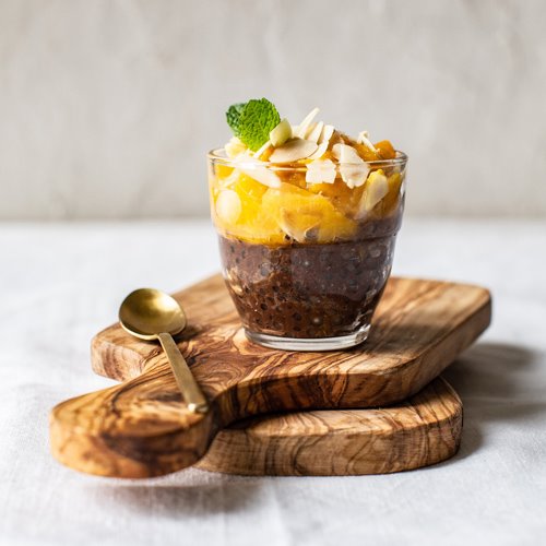 Deser chia z mango i płatkami migdałów, udekorowany listkiem mięty, podany w szklance na drewnianej desce z oliwkowego drewna, obok złota łyżeczka.