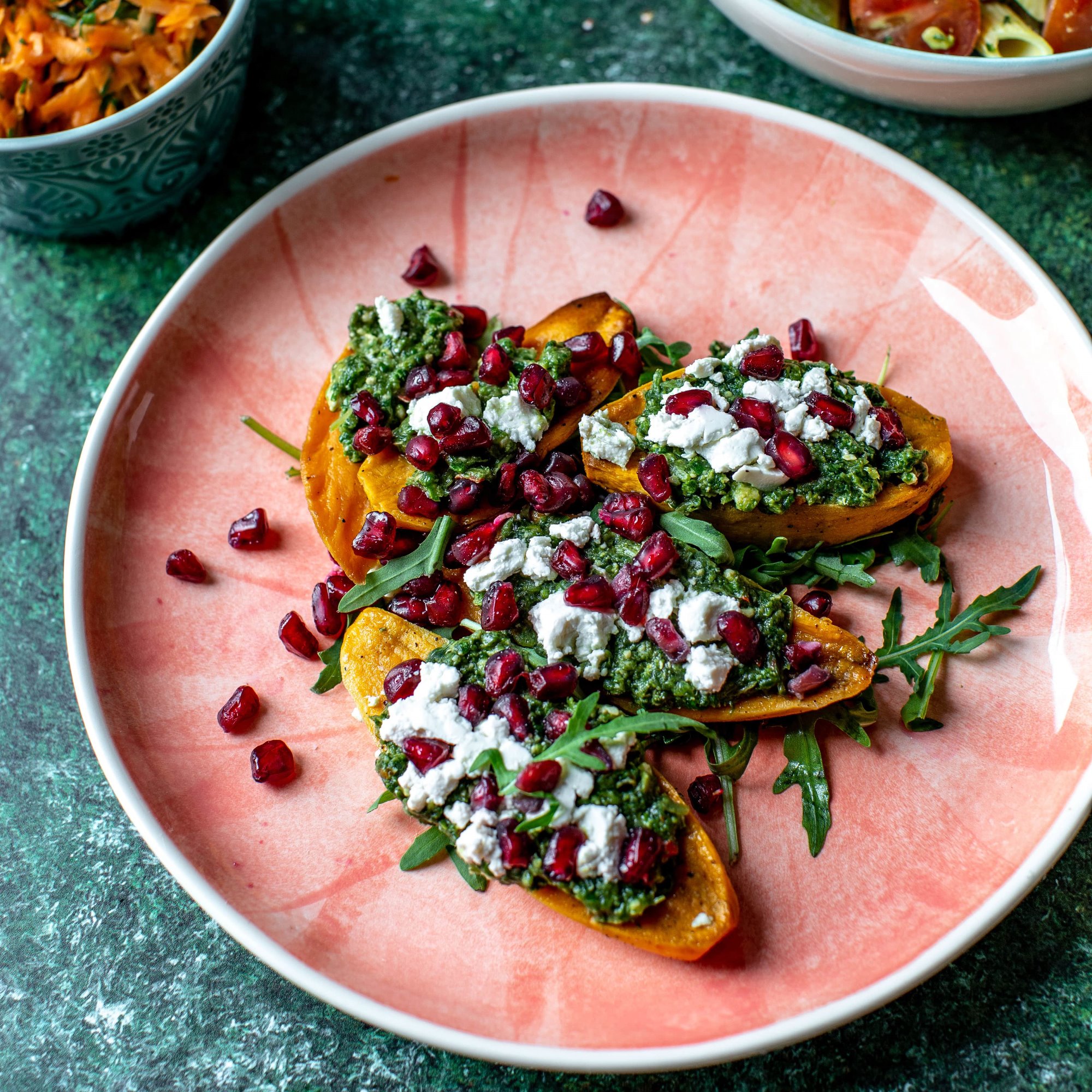 Pieczona dynia piżmowa z pesto, serem feta i granatem, podana na różowym talerzu z zielonym marmurkowym tłem, widoczne inne dania w miseczkach.
