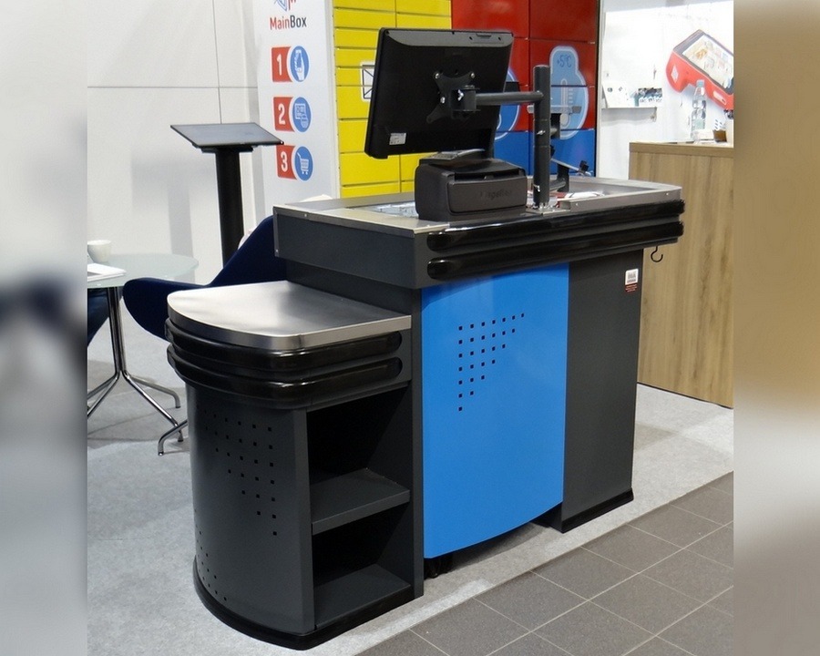 Stanowisko kasowe MINI w kolorach szaro-niebieskich z metalowym blatem i monitorem, widoczne na tle ekspozycji targowej z grafikami i elementami wyposażenia.