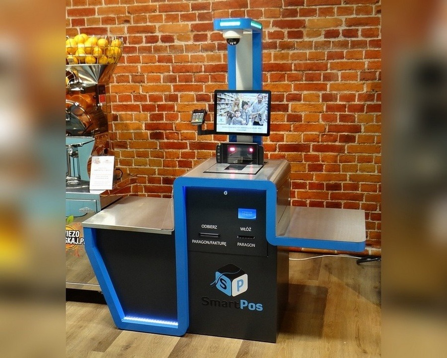Samoobsługowe stanowisko kasowe SmartPos z niebieskimi akcentami, wyświetlaczem pokazującym rodzinę i kamerą, na tle ściany z czerwonej cegły.