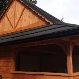 Drewniana altana z czarnym dachem z dachówki bitumicznej, widoczne rynny i dekoracyjne wykończenie szczytu dachu.