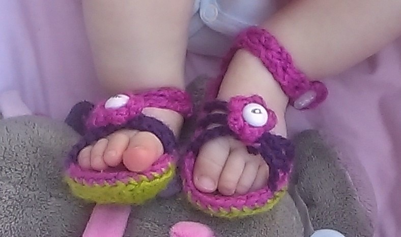 Ręcznie robione, szydełkowe buciki-sandałki w kolorach fioletu, różu i limonki na małych stópkach dziecka, z dekoracyjnymi kwiatkami i zapięciem na kostce.