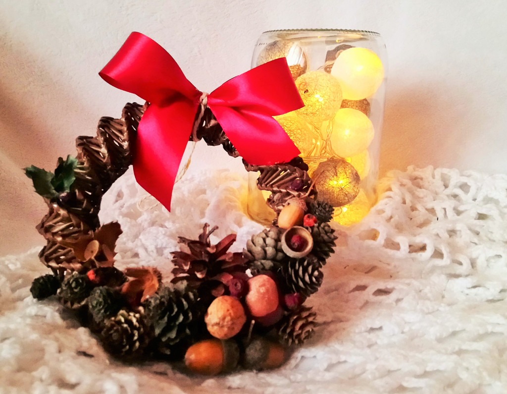 Dekoracyjny wieniec świąteczny z szyszkami, żołędziami i czerwoną kokardą, obok słoik z lampkami cotton balls, na białej, dzierganej serwecie.