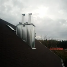 Nowe, błyszczące, metalowe obróbki kominowe i kominy wentylacyjne na dachu pokrytym brązowym gontem bitumicznym, widoczne okno dachowe i fragment drzew na tle pochmurnego nieba.