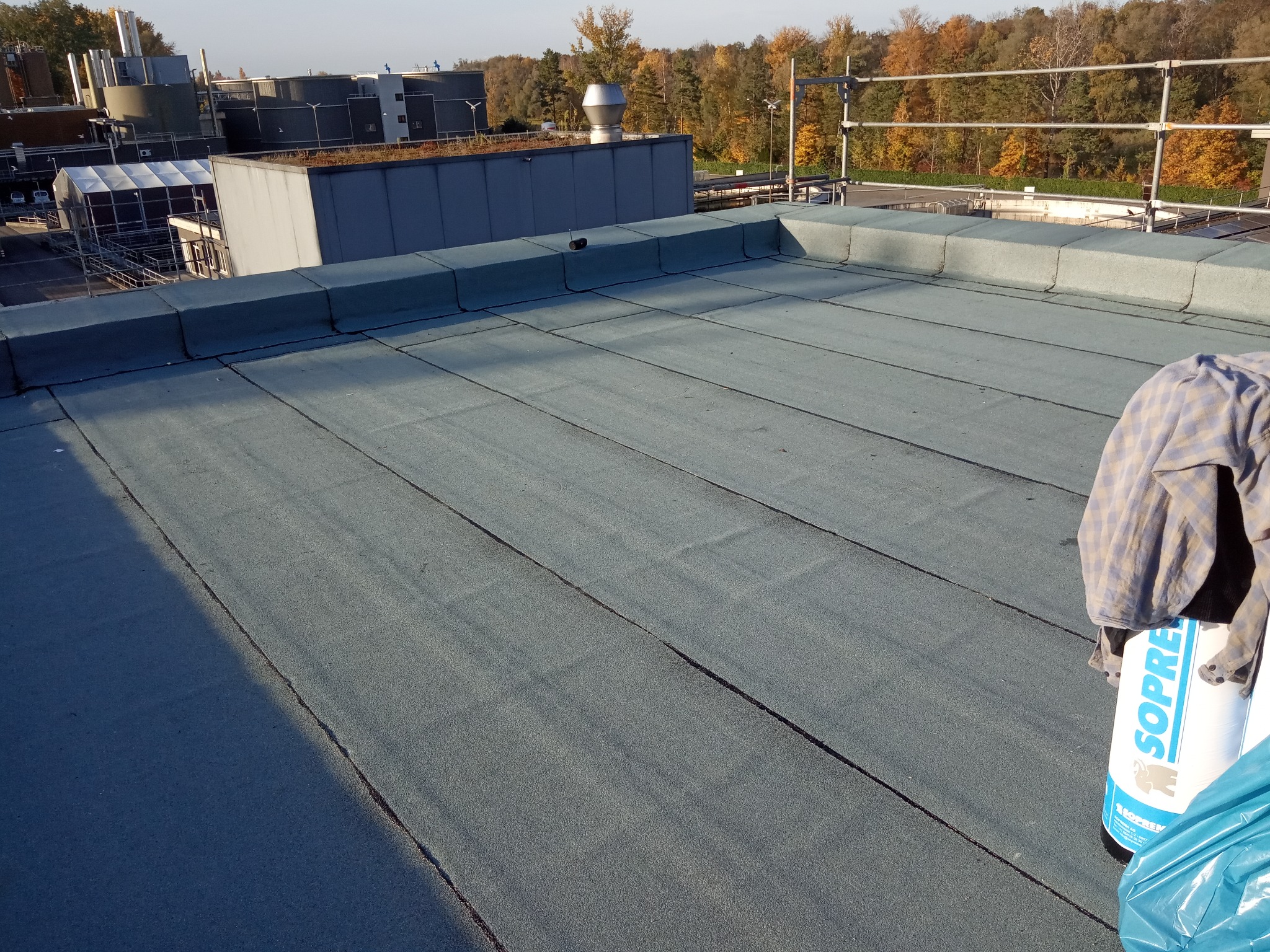 Płaski dach pokryty papą termozgrzewalną Soprema, widoczne zgrzewy, w tle przemysłowy budynek z zielonym dachem i jesienne drzewa.