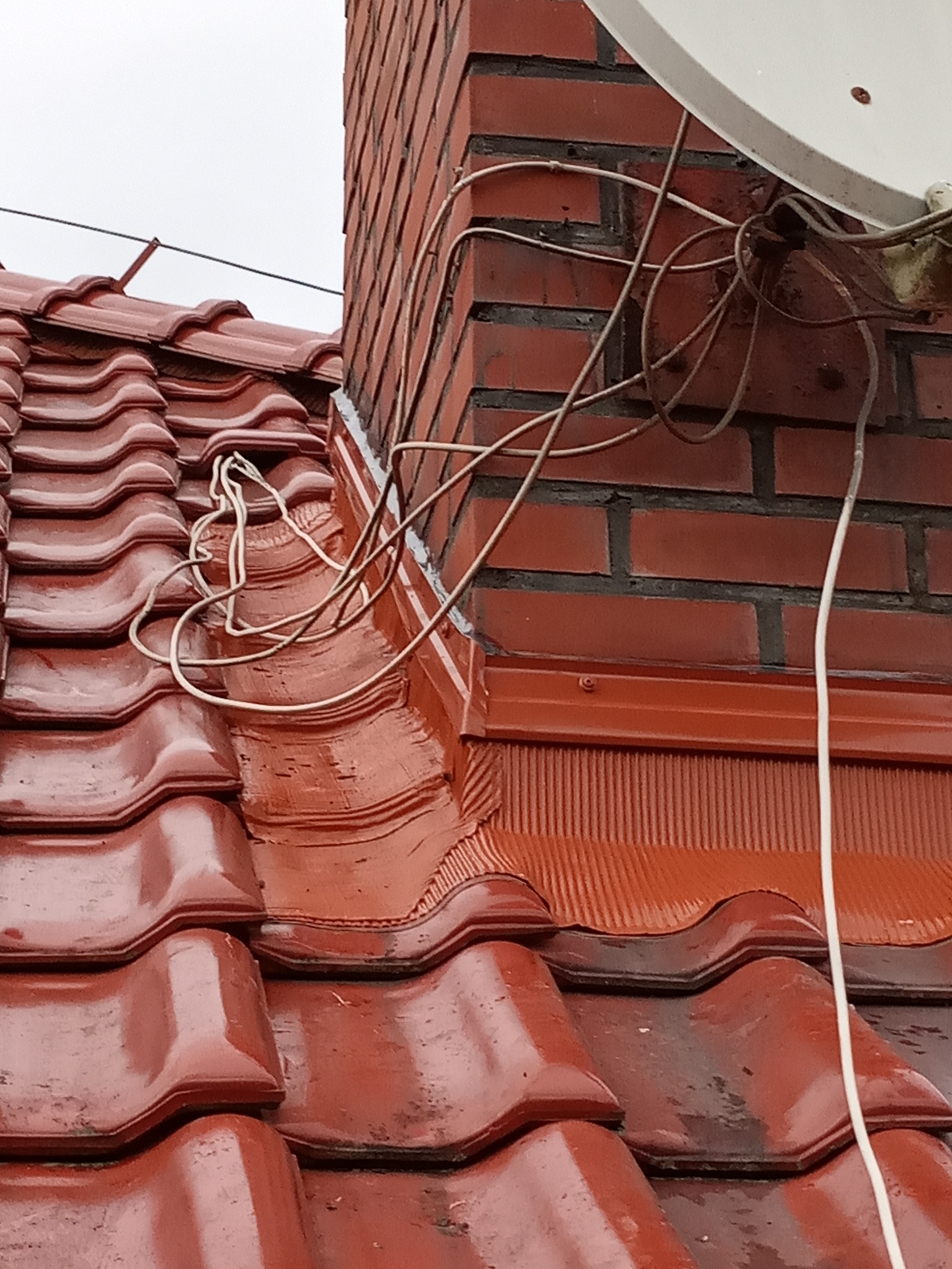 Czerwona dachówka ceramiczna, obróbka blacharska wokół komina z cegły, kable antenowe i antena satelitarna, pochmurne niebo.