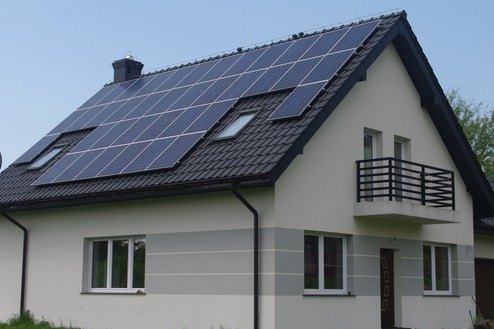 Dom jednorodzinny z panelami słonecznymi na dachu pokrytym czarną dachówką, widoczne okna dachowe i mały balkon z czarną balustradą.