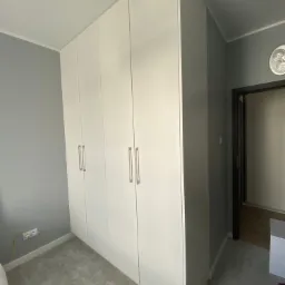 Biała szafa wnękowa z pięcioma drzwiami i prostokątnymi uchwytami, dopasowana do szarego wnętrza sypialni z widocznym gniazdkiem elektrycznym i fragmentem łóżka.