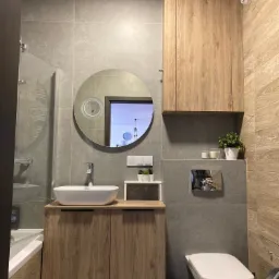 Nowoczesna łazienka z drewnopodobną zabudową umywalki i szafkami wiszącymi, okrągłe lustro z podświetleniem, szare płytki na ścianach i wisząca toaleta.